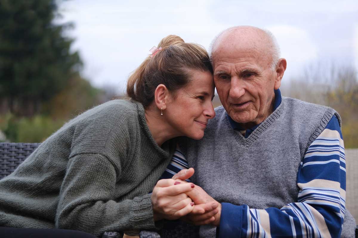 a woman holding hands with and sitting close to her senior father as they sit outside