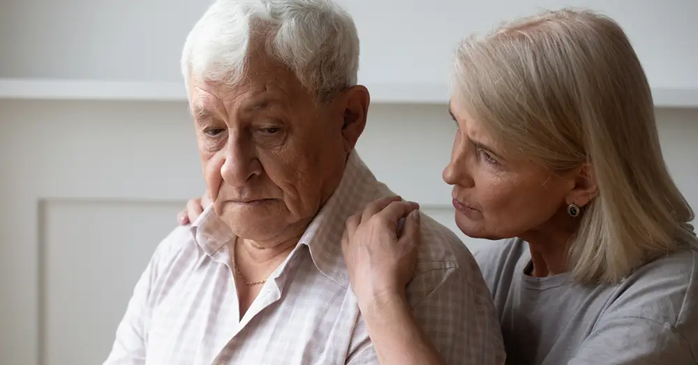 senior woman gently placing her arms around her stressed, senior husband